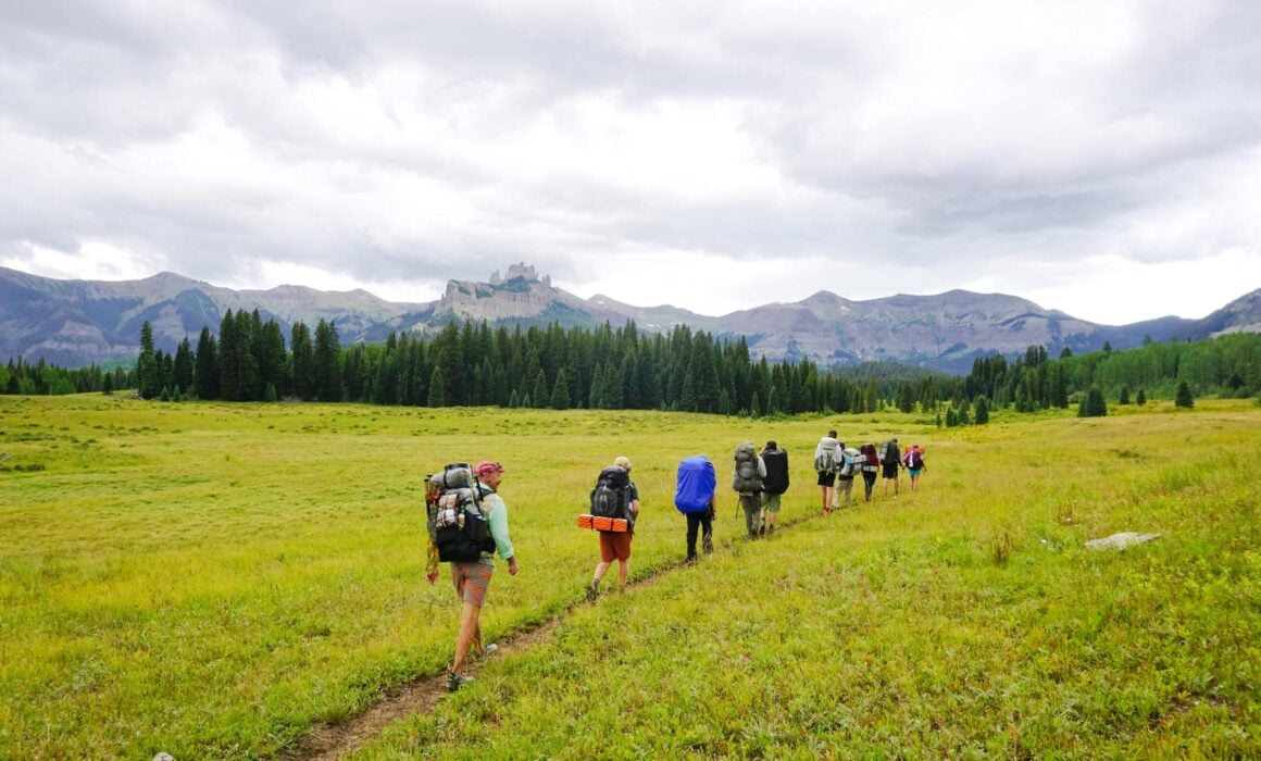 An ROE land-based class in single file backpacks up a flat mountain meadow with the Castles and West Elk Peaks in the background.