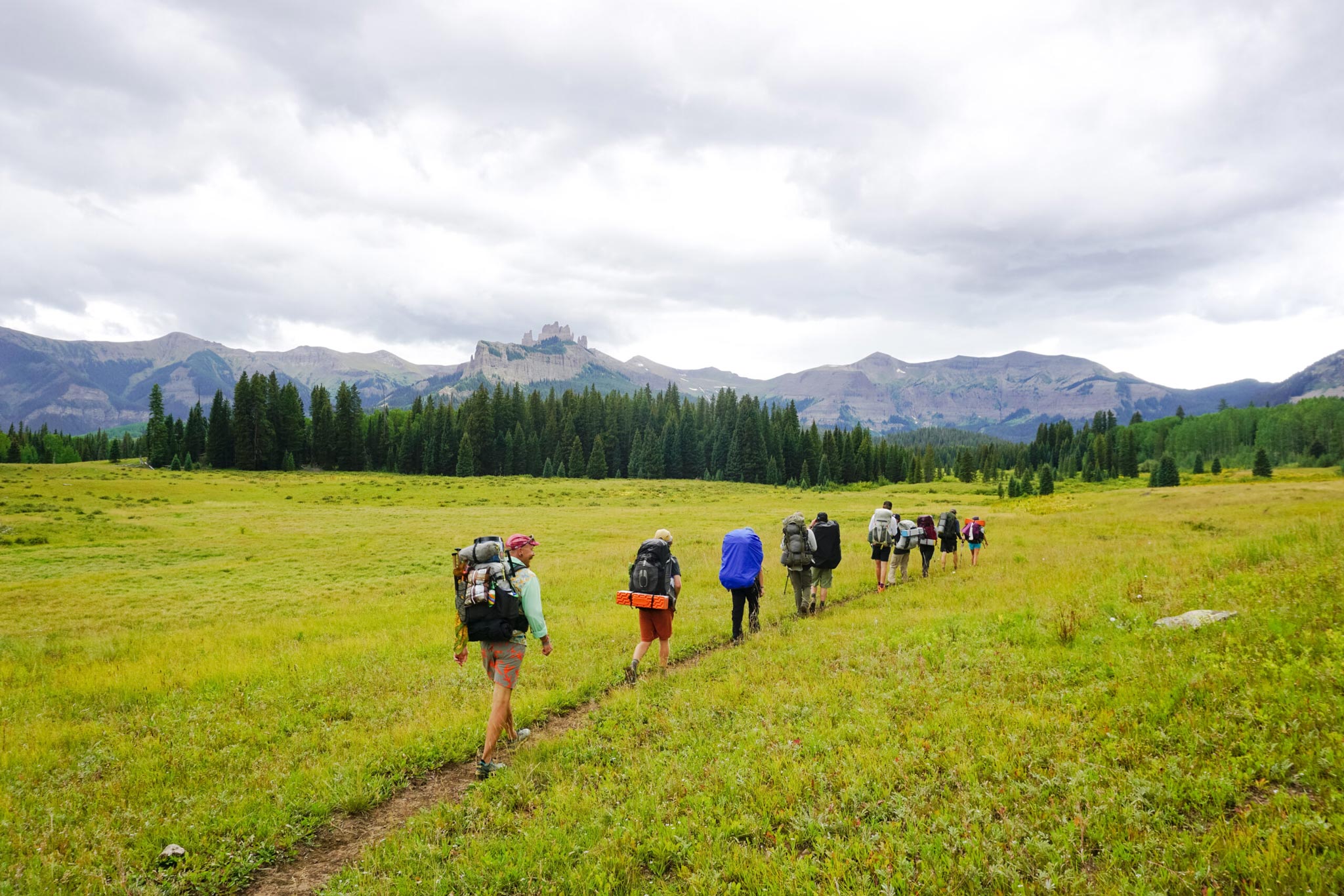 An ROE land-based class in single file backpacks up a flat mountain meadow with the Castles and West Elk Peaks in the background.
