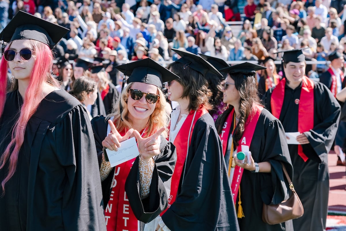 In a crowd of people, a graduate holds up a W with her hands.