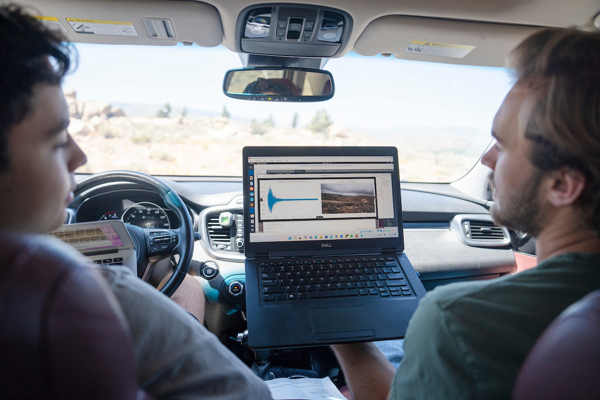 Two students look at a side by side comparison of a graph recorded by a sensor and a corresponding trail cam photo while sitting in a car a retrieving data from the devices which are tracking the traffic coming in and out of Hartman Rocks.