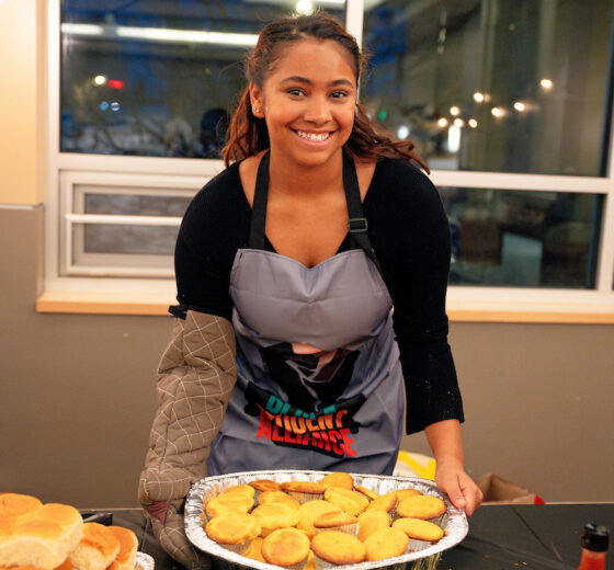 A student wearing an apron and oven mitt smiles while placing a tray of cornbread muffins onto the buffet table during Soul Food Night.