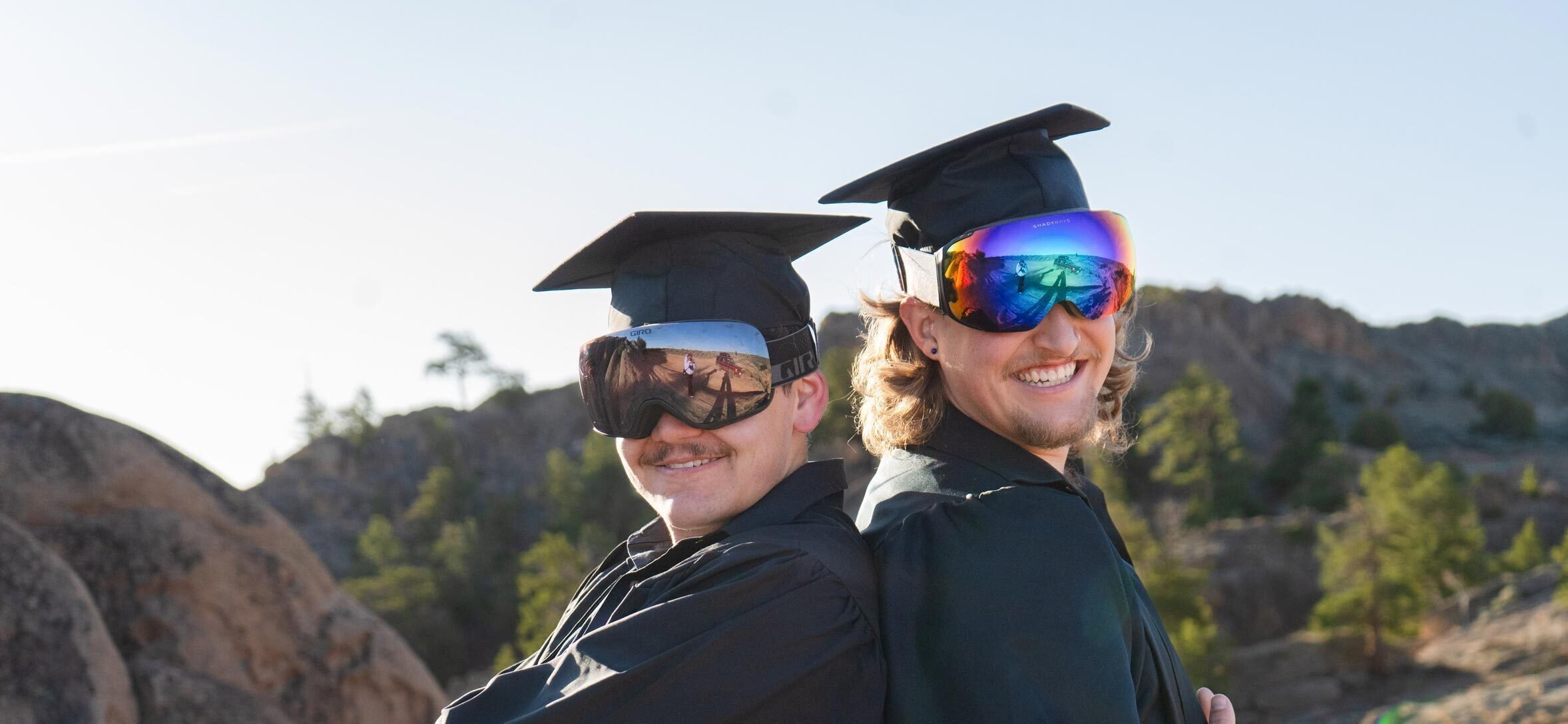 Two students wearing graduation caps, gowns, and ski goggles.