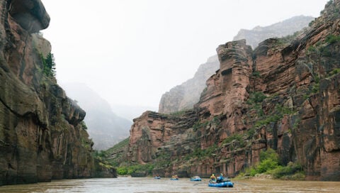 Rafters navigate a muddy river through a narrow canyon flanked by towering red rock cliffs under an overcast sky.