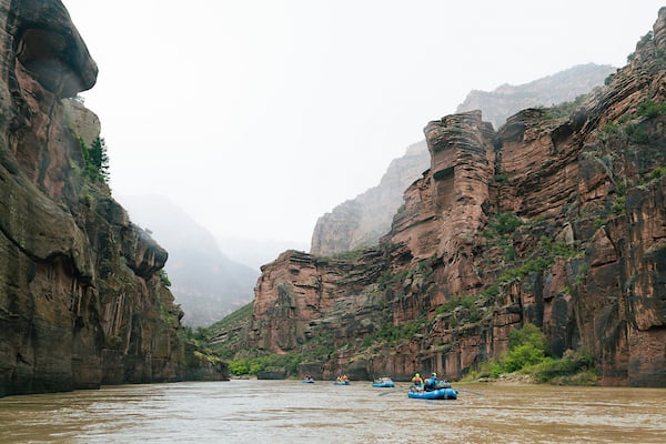 Rafters navigate a muddy river through a narrow canyon flanked by towering red rock cliffs under an overcast sky.