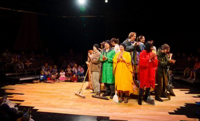 Student actors dressed in rain coats and ponchos stand on and around a wooden platform in the studio theatre as they sing together for the final scene of Western Theatre Co's production of 'LOVE'.