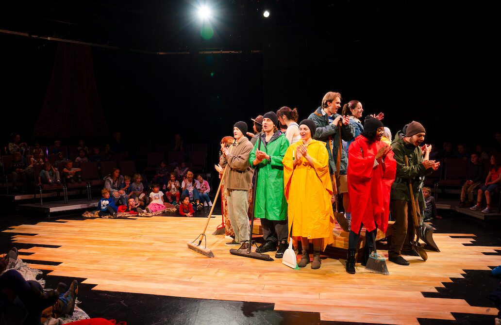 Student actors dressed in rain coats and ponchos stand on and around a wooden platform in the studio theatre as they sing together for the final scene of Western Theatre Co's production of 'LOVE'.