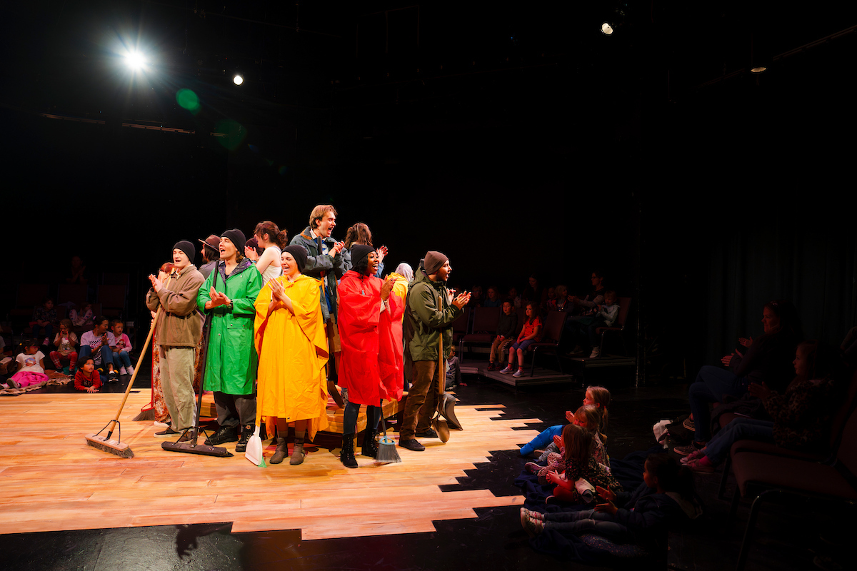 Student actors dressed in rain coats and ponchos stand on and around a wooden platform in the studio theatre as they sing together for the final scene of Western Theatre Co's production of 'LOVE'.