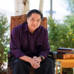 A person sits in a decorative chair outdoors under trees, with a small table beside them holding a stack of books.