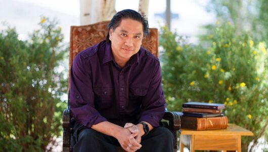 A person sits in a decorative chair outdoors under trees, with a small table beside them holding a stack of books.