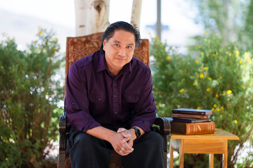 A person sits in a decorative chair outdoors under trees, with a small table beside them holding a stack of books.
