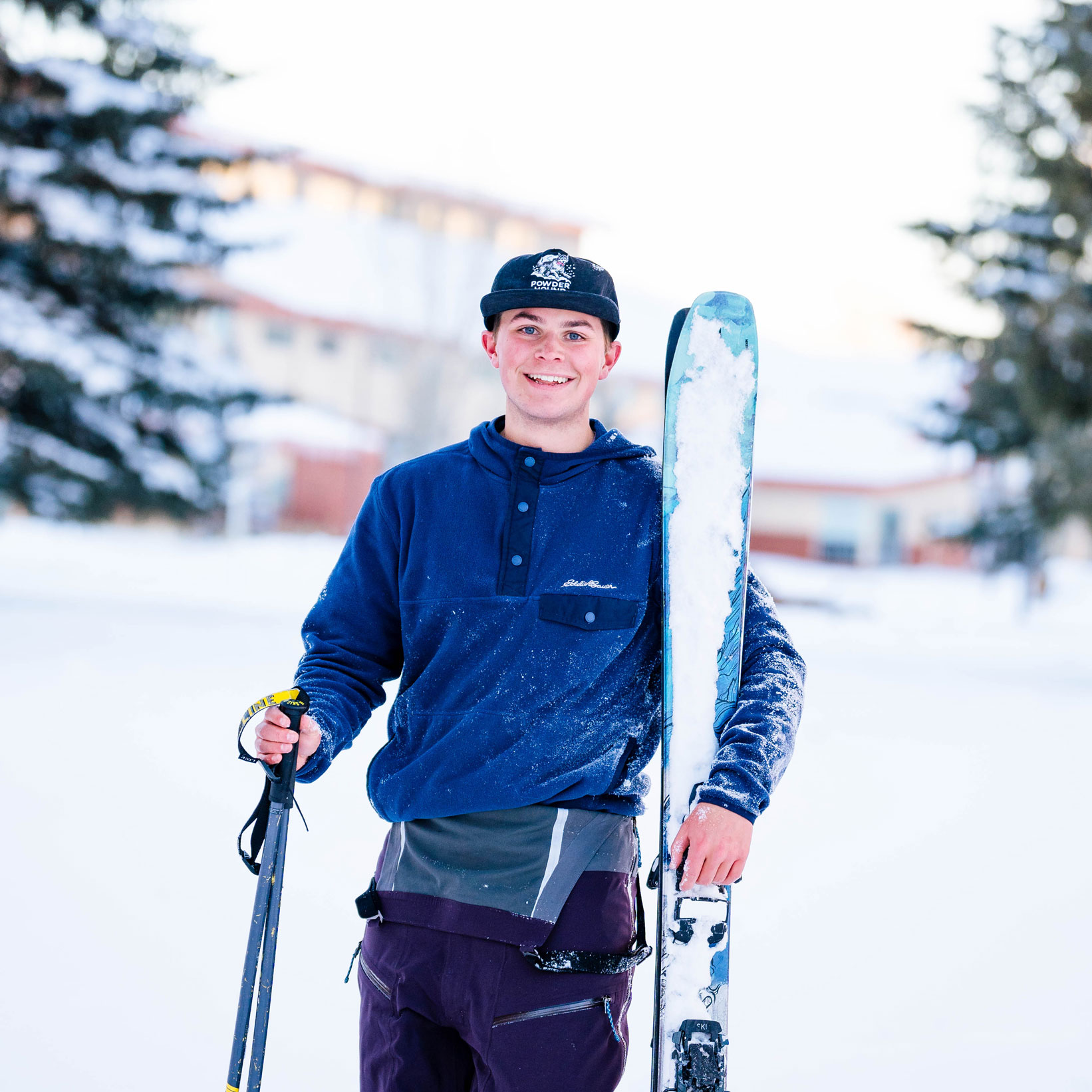 A student holds his skis and smiles with the Mountaineer Field House in the background.