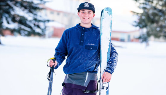 A student holds his skis and smiles with the Mountaineer Field House in the background.