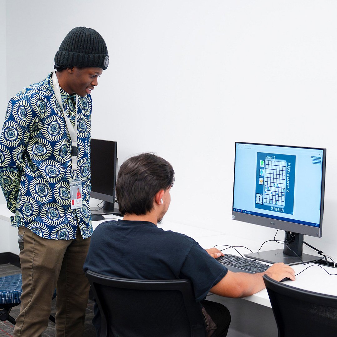 A student plays a game on a computer while another student watches over his shoulder during the CS club's game demo event.