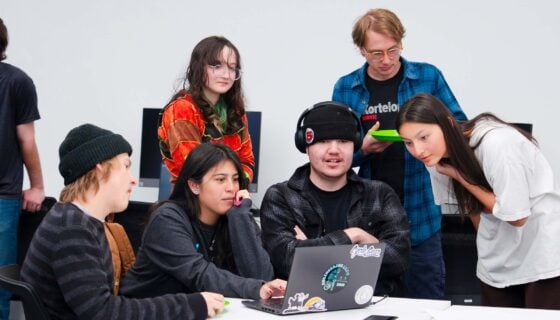 A goup of students gather around another student testing a game on a laptop at the CS club's game demo event.