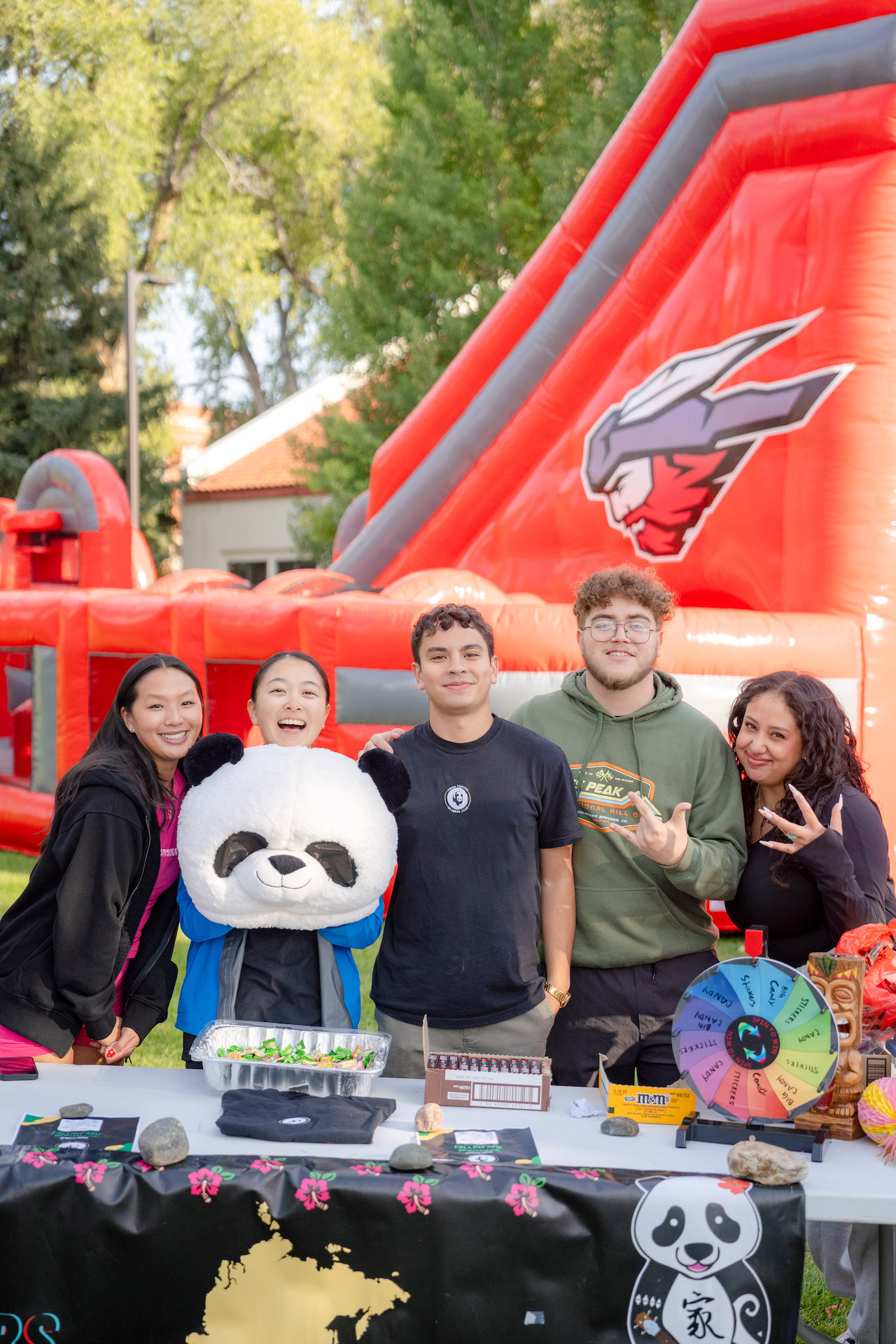 five APIC members smile at their booth at the club fair. (from left to right) female student smiles, female student holding a panda mask, male student smiles, male student smiles with a W, female student smiles and holds up a W