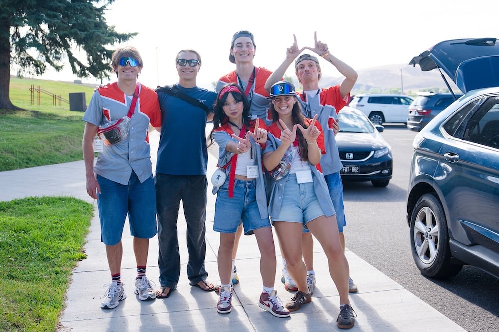 A group of six people in casual attire, some wearing red and gray outfits and accessories, pose cheerfully on a sidewalk near parked cars.