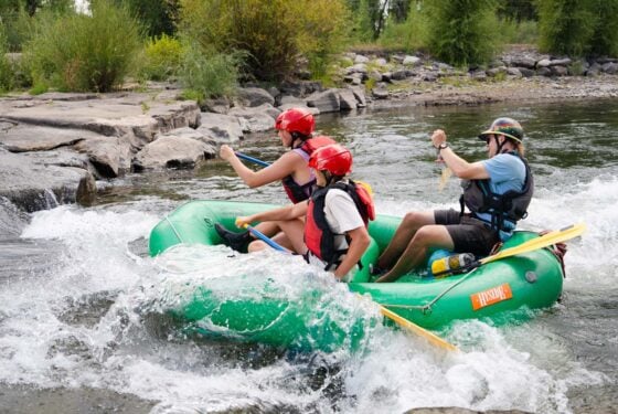 Three people in a green inflatable raft navigate whitewater rapids, paddling through turbulent waves with helmets and life vests.