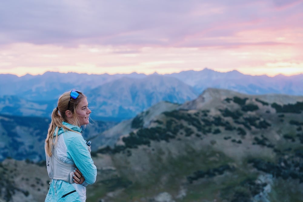 A person stands on a mountain ridge at sunset, gazing across a vast, layered landscape under a soft pink and purple sky.