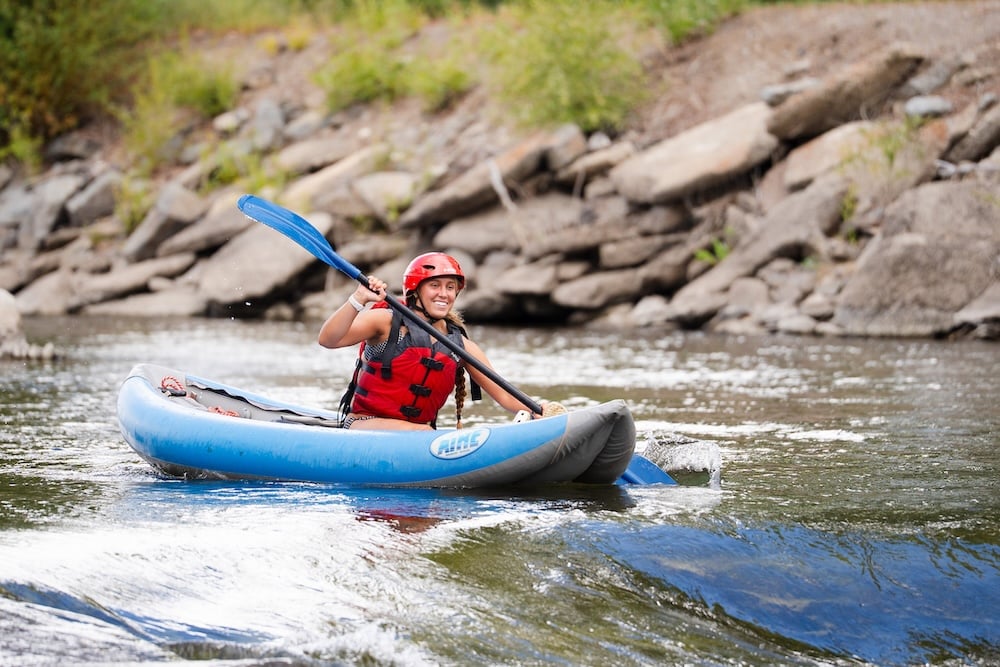 A person in a red helmet and life vest paddles a blue inflatable kayak through whitewater rapids near a rocky shoreline.