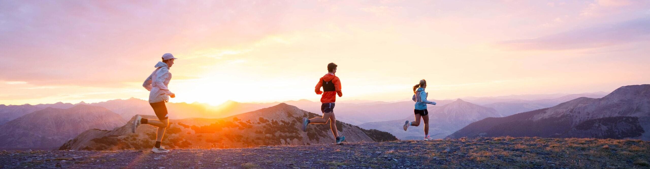 Three athletes run along a mountain ridge at sunrise.