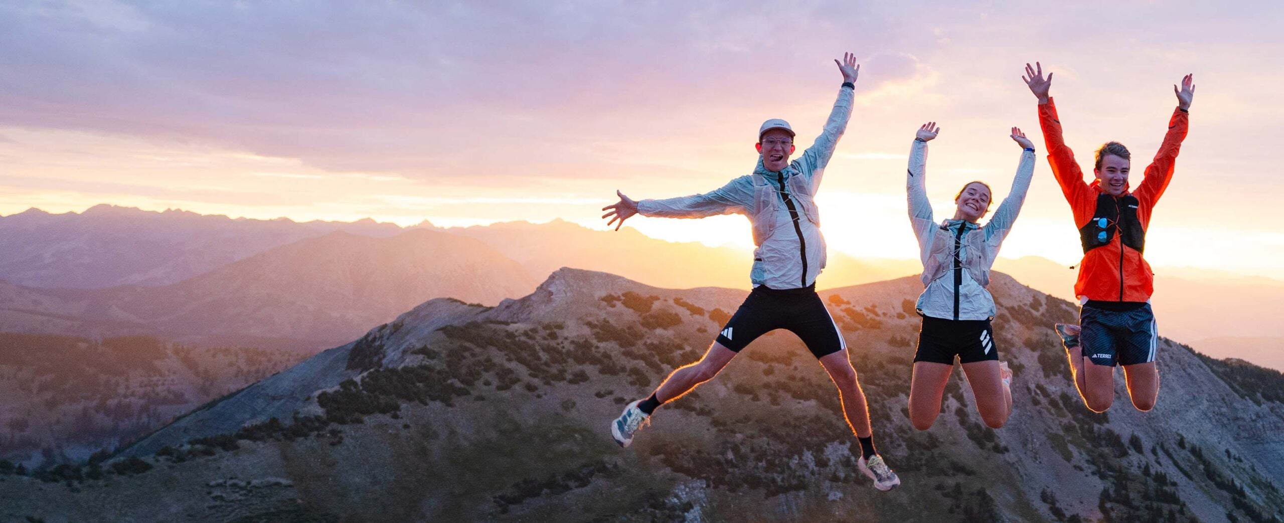Three trail runners jump on a mountain ridge with the sun rising behind them.