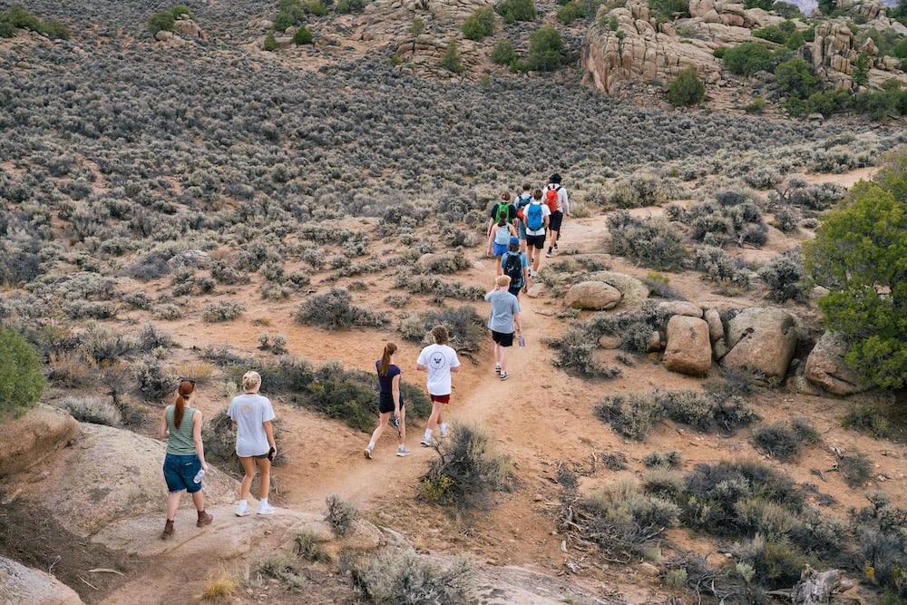 A group of hikers walks along a dirt trail through a desert landscape dotted with shrubs and large rocks.