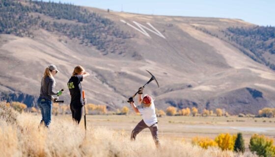 A student uses a pickaxe to dig a hole for sagebrush transplants. W Mountain can be seen in the background