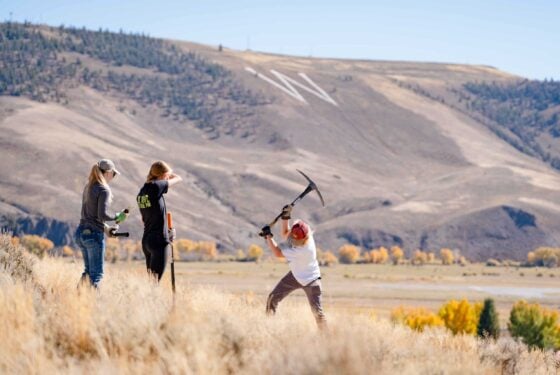 A student uses a pickaxe to dig a hole for sagebrush transplants. W Mountain can be seen in the background