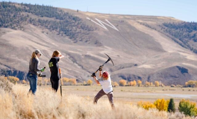 A student uses a pickaxe to dig a hole for sagebrush transplants. W Mountain can be seen in the background