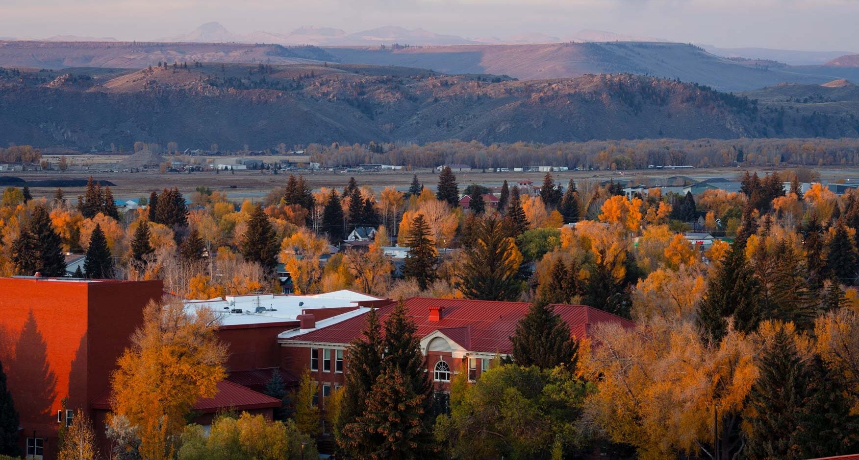 A scenic autumn view of a town nestled among trees with vibrant orange and yellow foliage, set against rolling hills and distant mountains under a soft, cloudy sky.