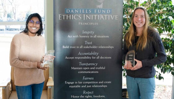 Two students, Kareena Hoover and Emma Wilke, hold the awards they won at the Daniel's Ethics Competition with the Daniels Fund Ethics Initiative Banner standing between them.