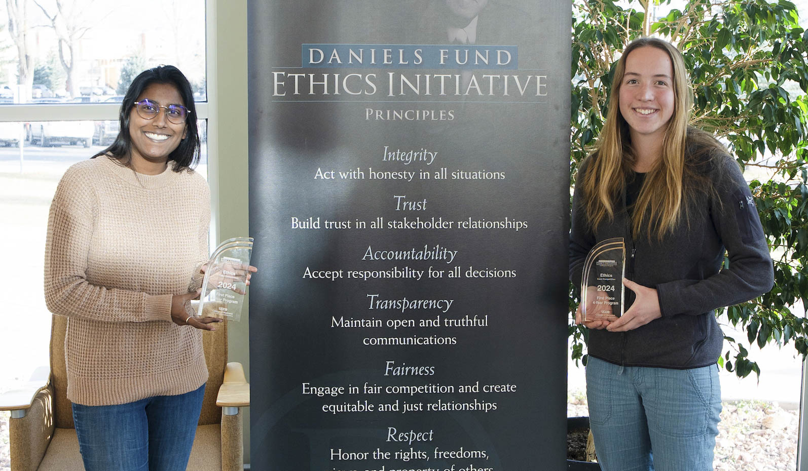 Two students, Kareena Hoover and Emma Wilke, hold the awards they won at the Daniel's Ethics Competition with the Daniels Fund Ethics Initiative Banner standing between them.