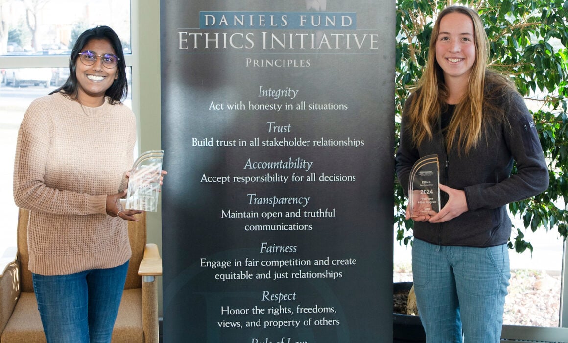 Two students, Kareena Hoover and Emma Wilke, hold the awards they won at the Daniel's Ethics Competition with the Daniels Fund Ethics Initiative Banner standing between them.