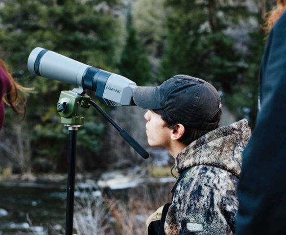 A student looks through a scope on a tripod at golden hour. Pine trees and Taylor River can be seen in the background.