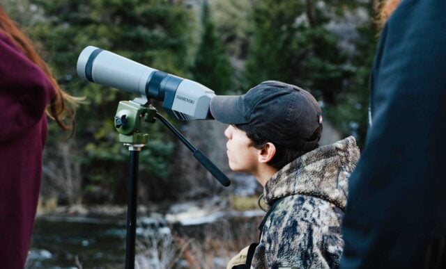 A student looks through a scope on a tripod at golden hour. Pine trees and Taylor River can be seen in the background.