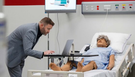 A man standing over a nursing dummy while working on his computer.