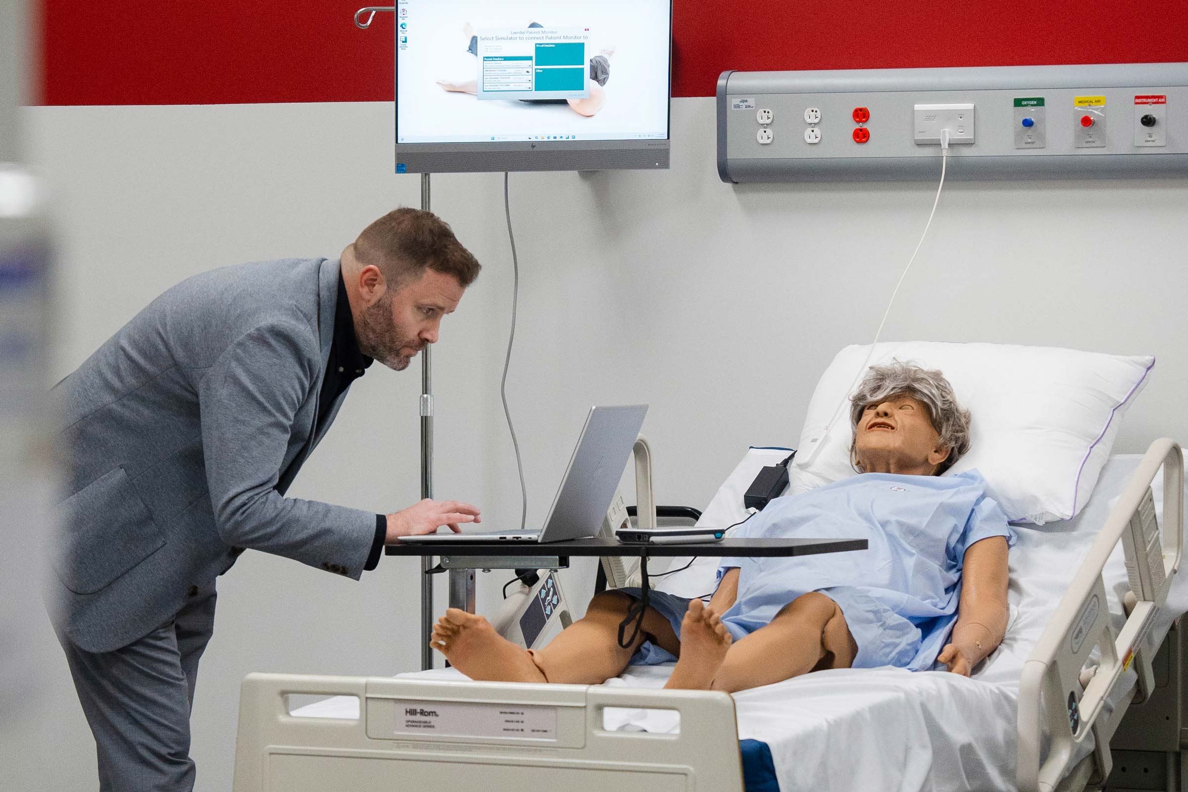 A man standing over a nursing dummy while working on his computer.