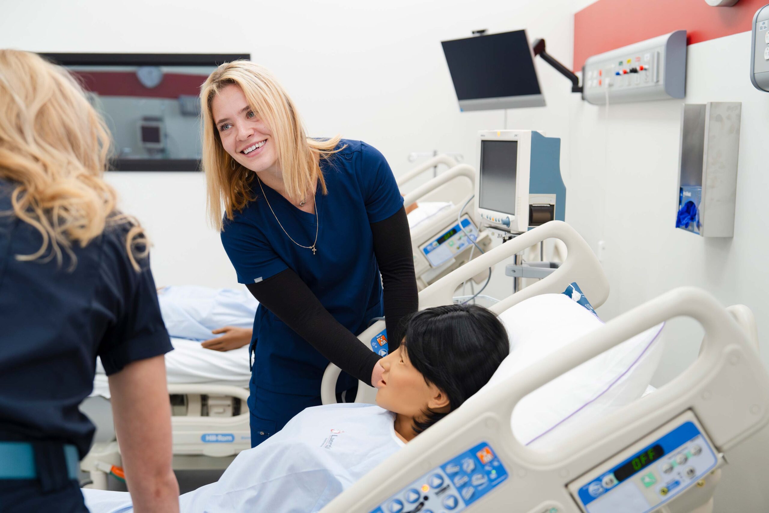 A nursing student smiles at her classmate while feeling for the pulse on a mannequin's neck during a simulation.