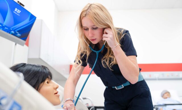 A nursing student uses a stethoscope to listen to a mannequin's breathing and pulse during a simulation.