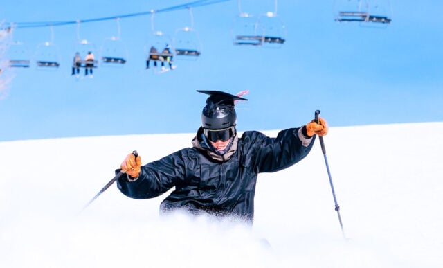 A student wearing a graduation cap and gown slashes his skis and picks up snow with a blue sky and chairlift in the background.