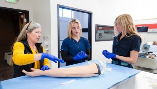 Two nursing students remove packaging from IV needles as their professor guides them on the correct procedure. A mannequin arm meant for practicing IV insertions lies on the table in front of them.