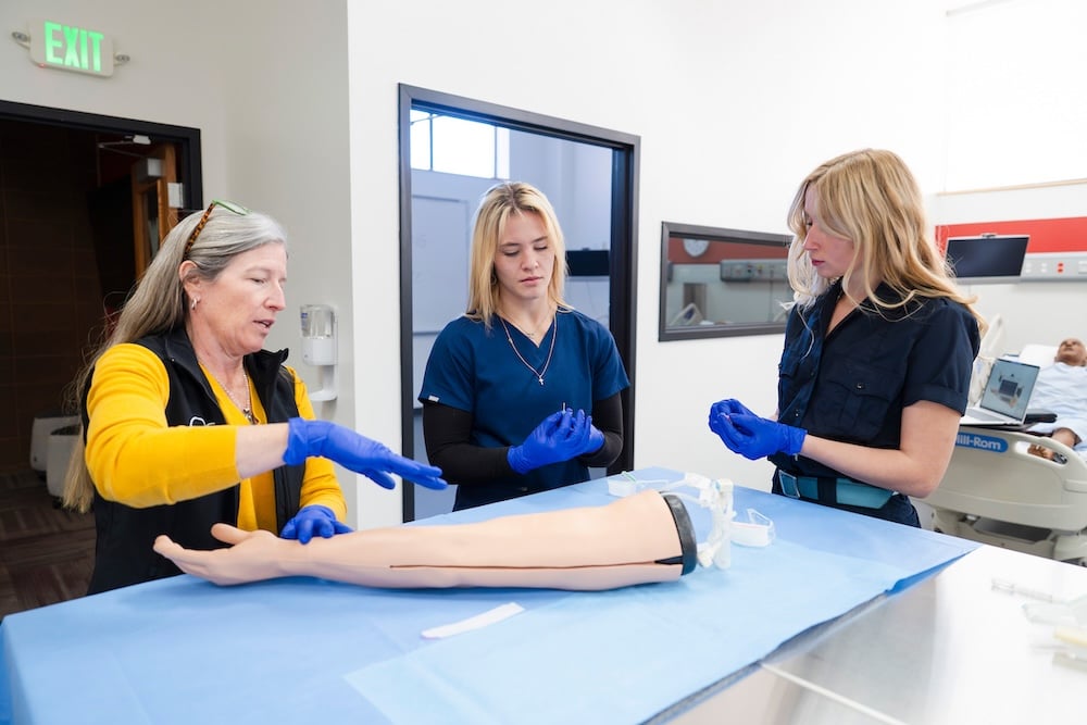 Two nursing students remove packaging from IV needles as their professor guides them on the correct procedure. A mannequin arm meant for practicing IV insertions lies on the table in front of them.