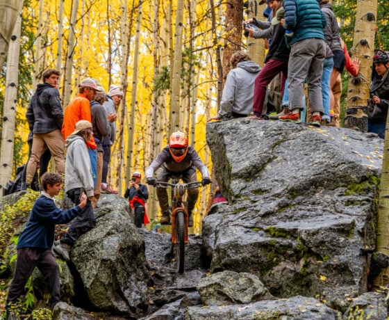 An athlete mountain bikes down a rock garden as students cheer around him during a race in the fall.