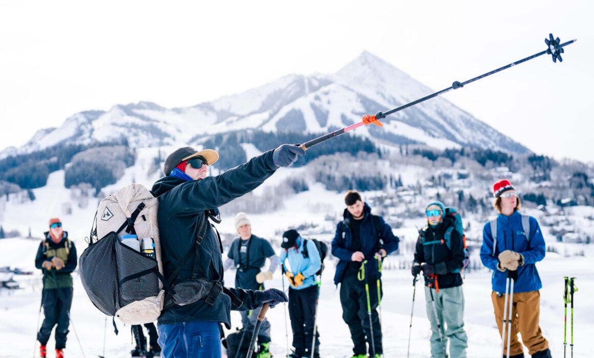 Professor Dave Erbe points his ski pole uphill in front of his backcountry skiing class. Mount Crested Butte is seen in the background.