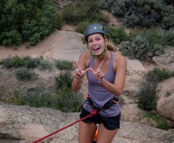 The student makes a W with her hands while she rock climbs.