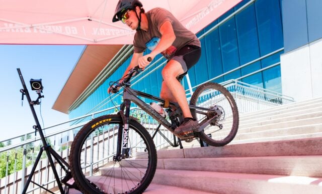 A student rides a mountain bike down the Rady Building stairs with a motion-sensing camera on a tripod behind him.