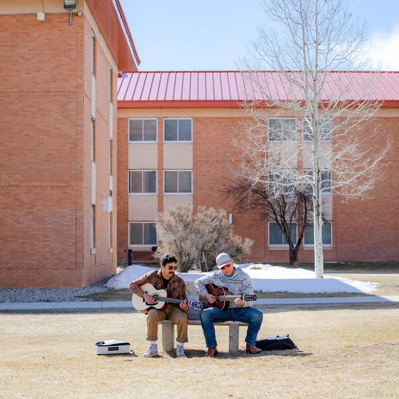 Two students sit on a bench and play guitar in front of the Mears Dorm Complex.