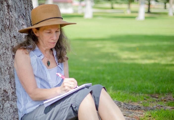 A creative writing student sits under a tree on Taylor Lawn and writes in her journal