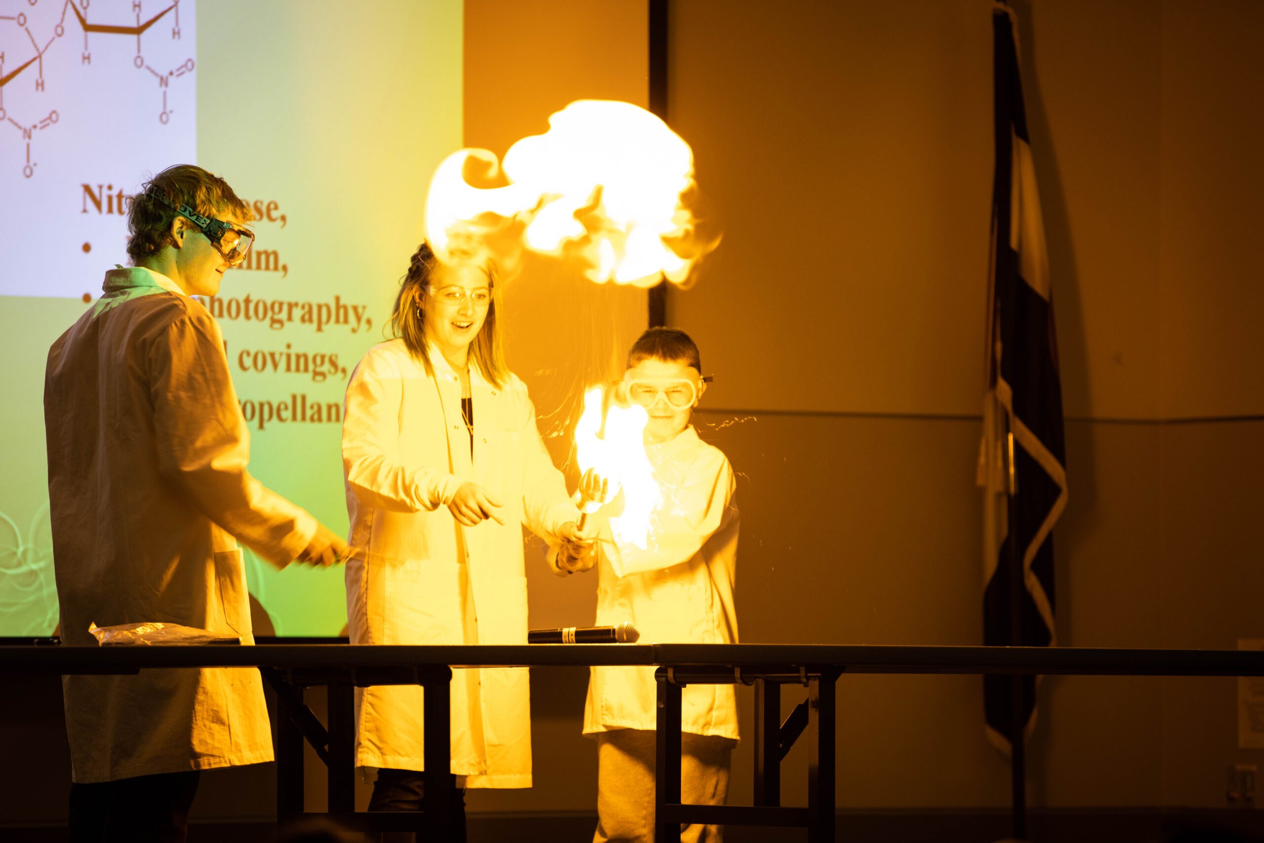 Two chemistry students and a little kid watch as they piece of nitrocellulose lights on fire in a burst of flames.