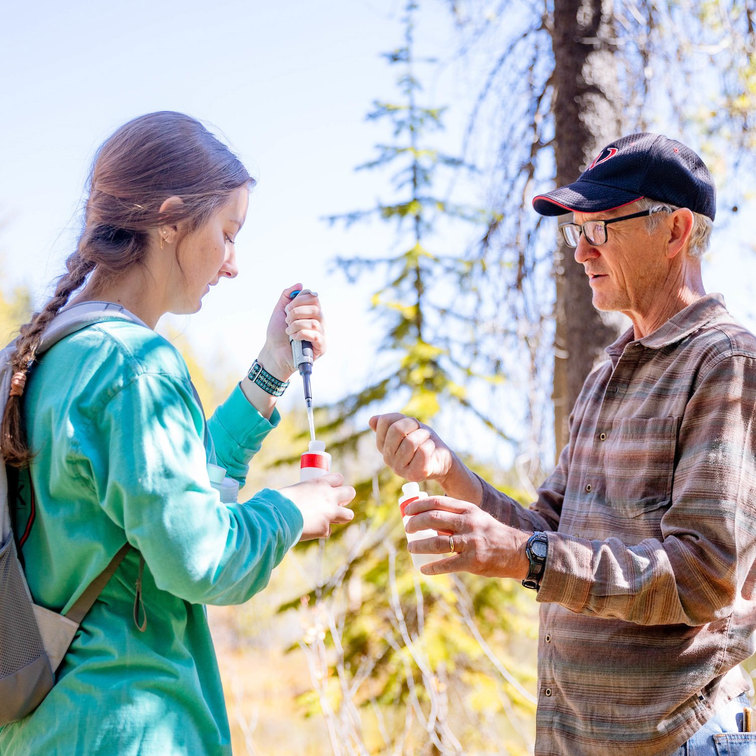 A professor demonstrates how to inject a water sample to a student as they stand outside in front of a pond.